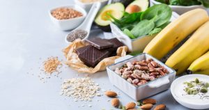 Lebensmittel wie Nüsse, Hülsenfrüchte, Avocado, Spinat, Bananen und dunkle Schokolade sind auf einem Tisch verteilt