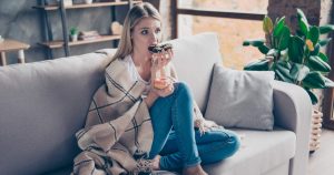 Frau sitzt in eine Decke gehüllt mit je einem Donut in der Hand auf dem Sofa und kämpft gegen Depressionen