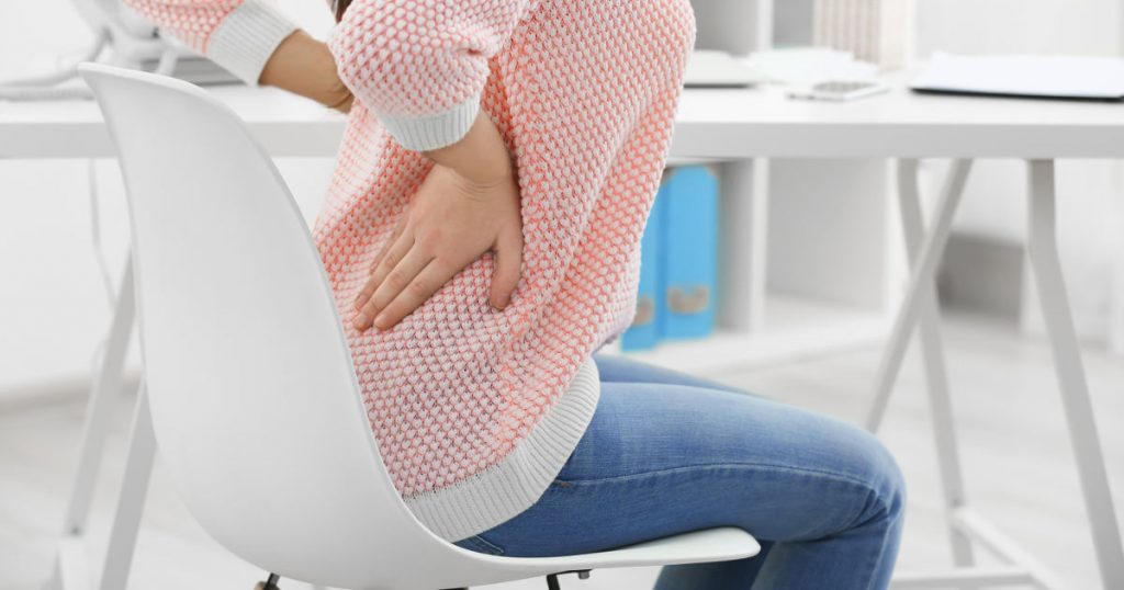 Frau sitzt auf einem Bürostuhl am Schreibtisch und hält sich vor Schmerzen den Rücken