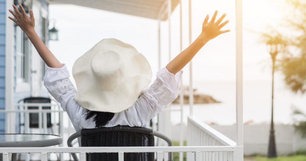 Frau mit Sonnenhut sitzt auf einem Balkon und streckt die Arme weit aus in Richtung Himmel und den letzten Sonnenstrahlen des Tages entgegen
