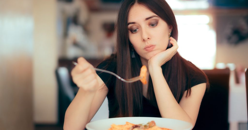 Zu sehen ist eine junge Frau, die ihr Essen, die Weizennudel auf der Gabel kritisch beäugt und hinterfragt, ob es gesund ist, diese zu essen