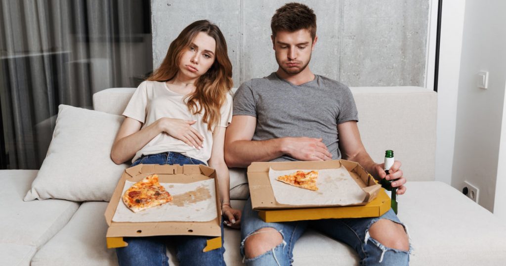 Ein unglückliches Paar sitzt mit halbleeren Pizzakartons auf der Couch und hält sich die vollen Bäuche
