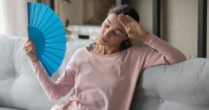 Eine Frau sitzt auf dem Sofa und fächelt sich mit einem Fächer Luft zu, da sie eine Hitzewallung hat