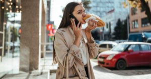 Eine beruflich beschäftigte Frau hält ihr Handy ans Ohr und beißt gleichzeitig in ein Sandwich