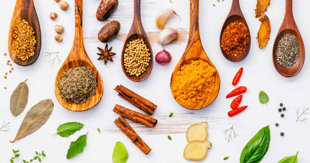 Es sind mehrere Gewürze auf Löffeln in Pulverform dargestellt. Daneben liegt jeweils das Gewürz in seiner natürlichen Form. Ingwerwurzel, Zimtstange, Chilischote, Lorbeerblatt