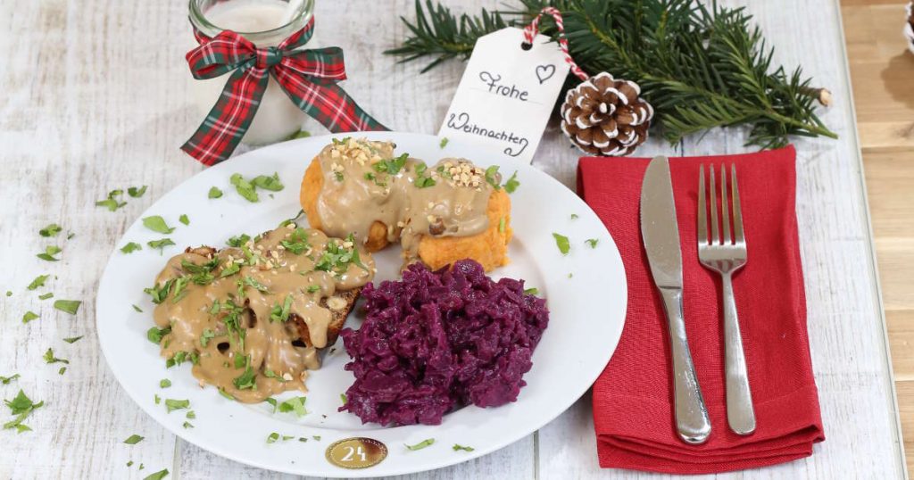 Ein Hauptgang mit Haselnussbraten, Süßkartoffelknödeln und Rotkohl ist schön auf einem Teller angerichtet. Daneben liegt Besteck und Weihnachtsdekoration