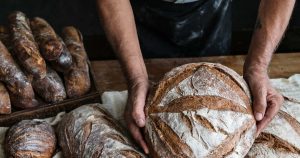 Ein Bäcker zeigt sein selbstgebackenes Brot. Neben ihm liegen einige Brotlaibe