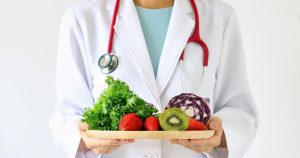 Eine Person im weißen Arztkittel und einem roten Stetoskop um den Hals hält in ihren Händen einen Holzteller mit pflanzlichen Lebensmitteln
