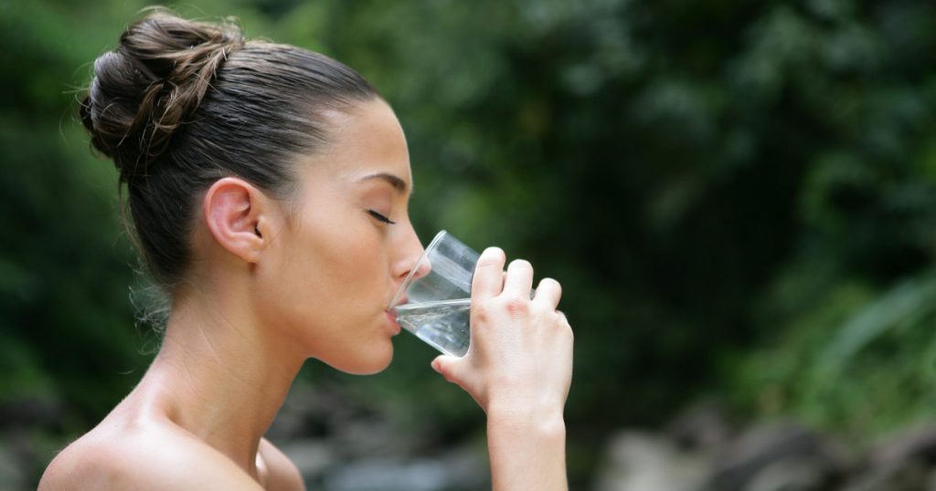 Eine junge Frau ist in Profilansicht abgebildet in der Natur und trinkt mit geschlossenen Augen ein Glas Wasser