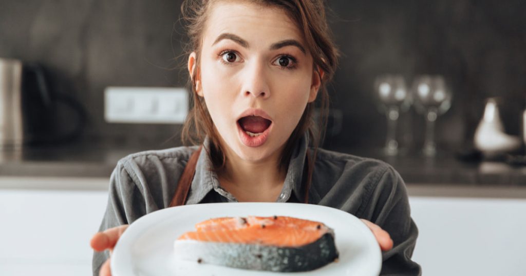 Eine junge Frau hält einen Teller mit einem Stück Lachs in den Händen. Sie guckt geschockt und hat den Mund weit aufgerissen.