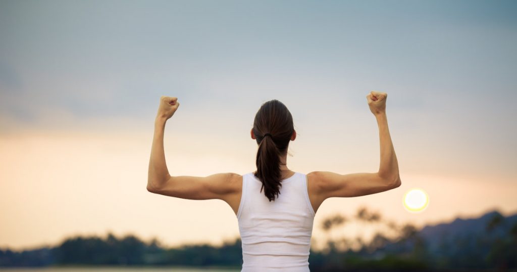 Eine durchtrainierte Frau steht vor einem Sonnenuntergang und spannt ihre Oberarme mit beidseitig gestreckten Armen an, um ihre Muskeln zu zeigen