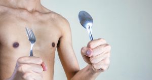 Der magere Oberkörper eines jungen Mannes ist zu sehen. Er hält Gabel und Löffel in jeweils einer Hand hoch und signalisiert damit, dass er essen und zunehmen möchte.