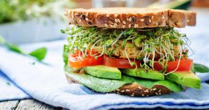 Ein Sandwich mit Sprossen, Avocado, viel Gemüse, einem Haferbratling und Vollkornbrotscheiben ist dargestellt.