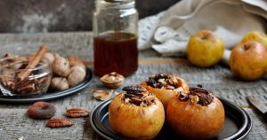 Drei gefüllte Bratäpfel mit Nüssen und Honig liegen im Vordergrund auf einem Teller. Im Hintergrund ist ein Glas Punsch zu sehen und daneben ein Teller mit Walnüssen.
