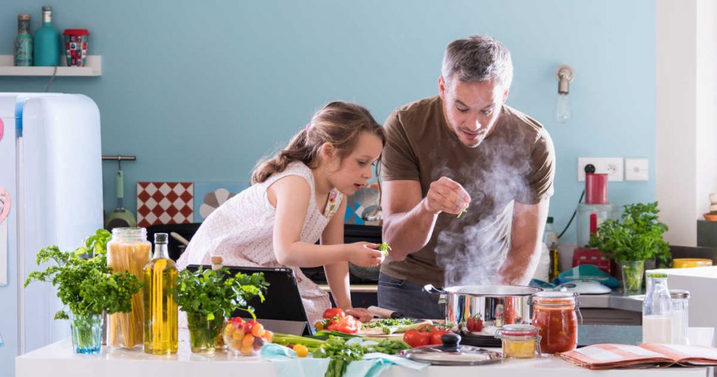 Ein Vater steht mit seiner Tochter zusammen am Herd und sie kochen gemeinsam. Um sie herum stehen viele frische Lebensmittel verteilt