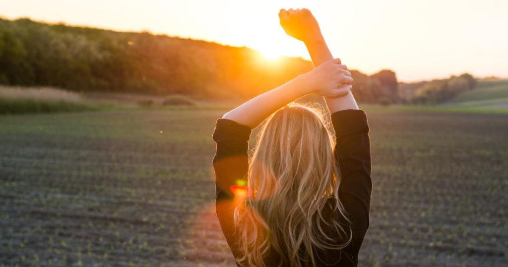 Eine junge Frau reckt die Arme empor und steht einem Sonnenaufgang gegenüber auf einer Wiese