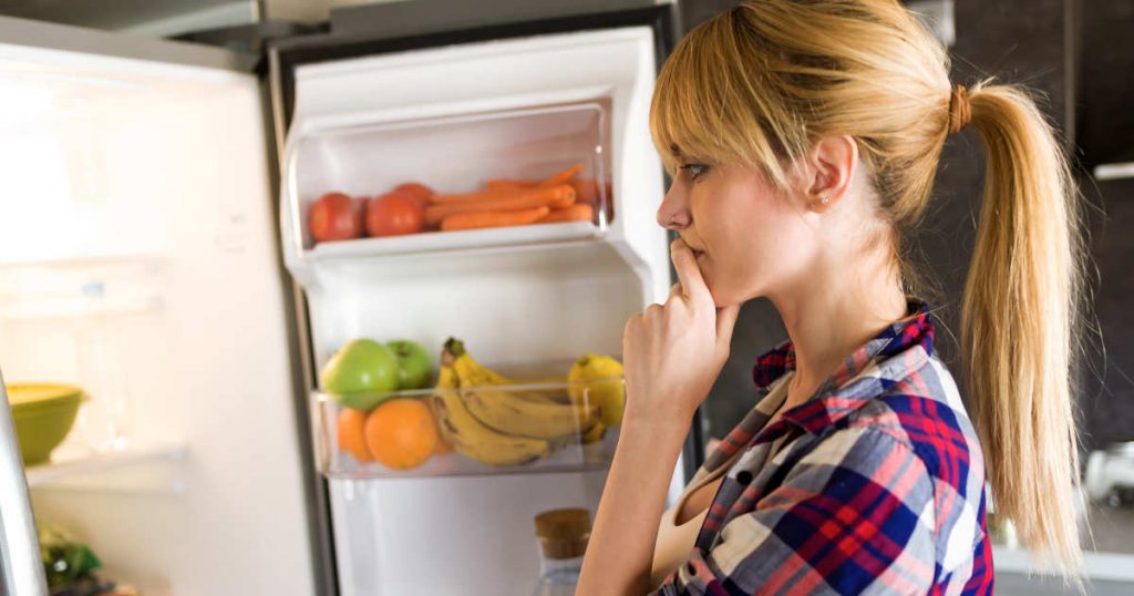 Eine Frau mit Pferdeschwanz steht vor dem offenen Kühlschrank, der mit buntem Obst und Gemüse gefüllt ist. Sie überlegt, was sie essen soll.